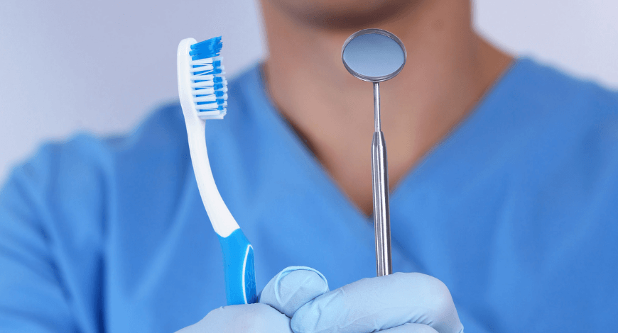Dr. Varajti Artúr fogorvos, szájsebész bal kezében egy fogászati tükröt, jobb kezében pedig egy fogkefét tart.