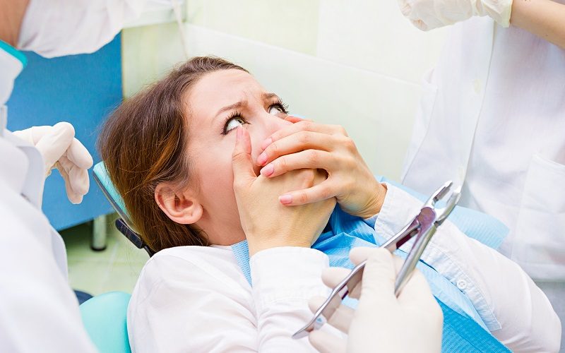 Egy fiatal nő ül a fogorvosi székben és fogja a száját, fél a fogorvosi vizsgálattól.