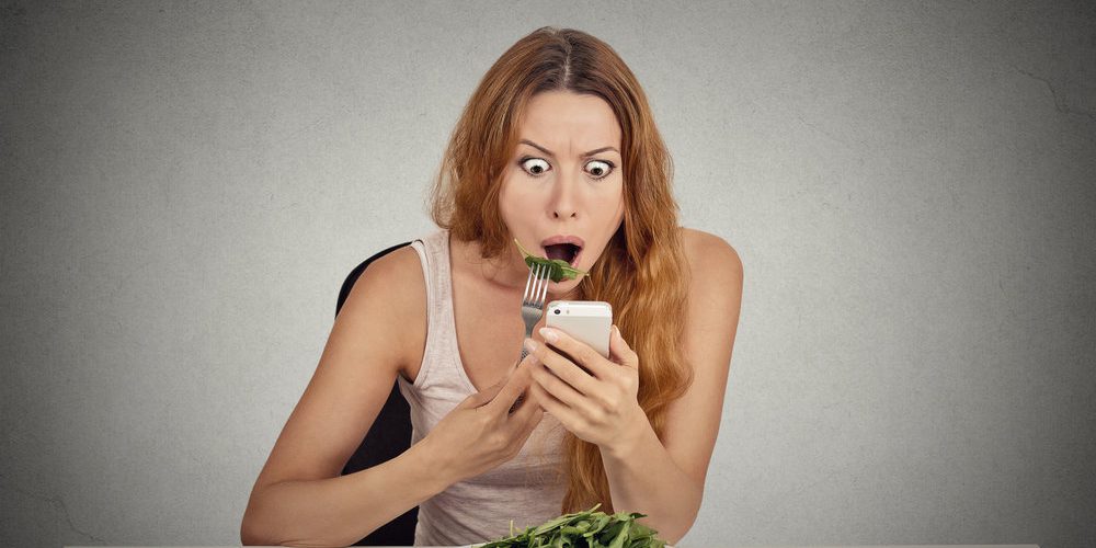 Vörös hajú nő eszik egy salátát, telefonon rossz híreket ad és döbbent arcot vág.
