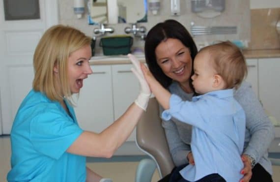 Dr. Friedreich Dóra elégedett, mosolygó páciens ölében tartott gyermekkel pacsizik az Artdent Fogászat rendelőjében
